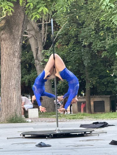 The image displays two individuals engaged in a synchronized pole acrobatics performance outdoors in a public urban green space or park area in Varna, Bulgaria. Both performers, appearing to be women, are inverted and suspended from a portable, silver-colored pole. They are positioned facing each other, with their legs wrapped around the pole for support, and their heads directed downwards. Each performer wears a matching long-sleeved, royal blue bodysuit adorned with a pattern of small, reflective white elements.

The pole is mounted on a circular black base with four extendable stabilizing arms spread across the grey paved ground. To the left of the pole's base, a pair of dark-colored slide sandals rests on the paving. Another partially visible dark slide sandal is present on the far right. A black soft bag, possibly a duffel bag, lies flat on the ground to the right of the pole base.

In the mid-ground, behind the performers and pole, a large, rough-barked tree trunk is prominent on the left. Further back, a person in a light-colored t-shirt and shorts is seated on a low, light-grey concrete bench near the base of another tree. The background is dominated by dense green foliage from numerous mature trees. To the right of the pole, behind the performers, a low, light-colored concrete structure with dark rectangular openings is visible. Adjacent to this structure, a bright red sculpture shaped like a hand making a "rock on" gesture stands vertically. The lighting suggests daytime, with diffused illumination across the scene.