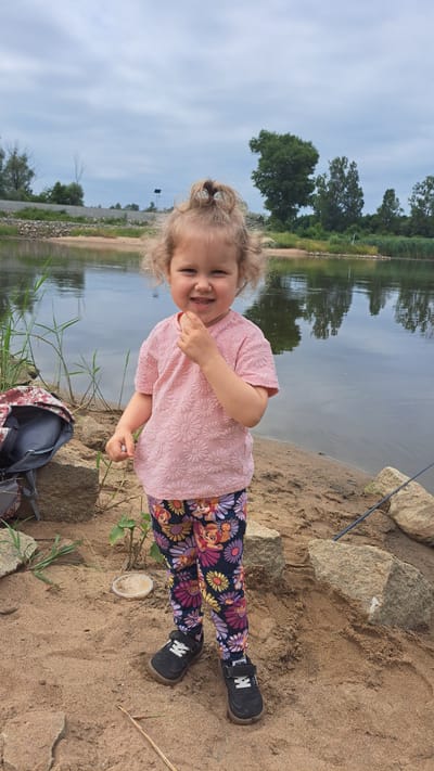 A young child, appearing to be a girl with blonde curly hair pulled into a top knot, stands on a sandy riverbank. She is wearing a pink textured t-shirt and patterned leggings featuring flowers and cartoon characters. The child is smiling and has her hand near her mouth. To her left, a backpack is visible. Behind her, a calm body of water reflects the overcast sky and trees lining the opposite bank. The environment suggests a natural setting, likely a park or recreational area near a river. There is no indication of the specific location within Krosno Odrzańskie, Poland.