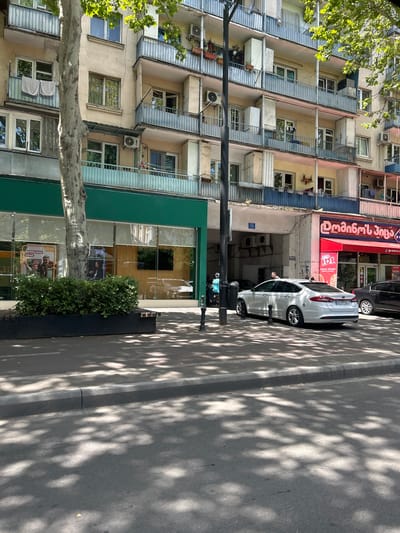 The image shows a street scene in Tbilisi, Georgia. In the foreground, a white sedan is parked on the side of the road, casting a shadow on the asphalt. The car has the license plate LC 679 LL. Behind the car, a building with multiple floors and balconies is visible. Some balconies have laundry hanging, and there are air conditioning units attached to the building's facade. Green bushes and a large tree with dappled sunlight are present on the sidewalk. To the right of the building, a red awning with Georgian text and a logo suggests a business, possibly a restaurant. Another car, a dark sedan, is parked further down the street. The overall impression is a typical urban streetscape with residential and commercial elements.