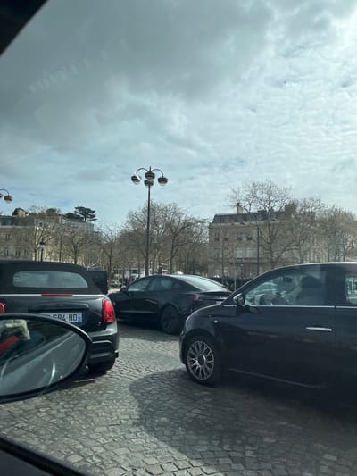 The image depicts an urban traffic scene on a cobblestone (pavé) street within Place de la Concorde, Paris, France. The perspective is from inside a vehicle, looking forward.
In the immediate foreground, the lower left shows the reflection in a side mirror and the rear of a black Mini Cooper Convertible with a French license plate (984 AD) partially visible. To the right of the Mini Cooper, a black Tesla Model 3 is positioned, seen from the rear. Further to the right and slightly in front, a dark-colored Fiat 500 is visible from its left side; a person with a light-colored head covering is discernible in the passenger seat. Vehicles are static or moving slowly, indicating traffic.
The background features typical Parisian Haussmannian architecture, including large, light-colored stone buildings with classical facades and mansard roofs. Between the street and these buildings lies a wide expanse of park-like area populated by numerous deciduous trees, currently bare. Ornate black lampposts with four globes are scattered throughout the scene. The sky is overcast with patchy clouds, and diffused sunlight creates soft shadows. Reflections from the viewing vehicle's interior are visible across the upper portion of the image.