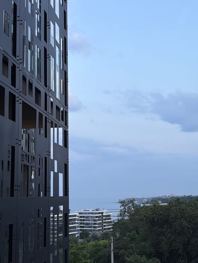 This media file captures an outdoor scene with a prominent architectural feature in the foreground. The foreground is dominated by a dark, perforated metal facade, possibly a building's exterior or a decorative screen. This facade is composed of many rectangular and square openings of varying sizes, some of which reveal glimpses of glass or reflective surfaces behind them, suggesting windows or panels. The metal is secured with visible rivets, adding to its industrial or modern aesthetic.
Beyond the metal structure, the background reveals a cityscape and natural elements. A tall, modern white building with a distinctive layered or stepped facade stands to the right, partially obscured by lush green trees. Further in the distance, a hilly landscape is visible, with more buildings, some appearing to have golden domes, and possibly cranes, suggesting ongoing construction or a developed urban area along a coastline. A body of water, likely the sea, can be seen in the middle ground. The sky above is a pale blue with scattered white clouds, indicating fair weather and likely daytime. The orientation of the image suggests it was taken during the day. The overall impression is one of contemporary architecture set against a backdrop of a city by the sea and natural greenery.