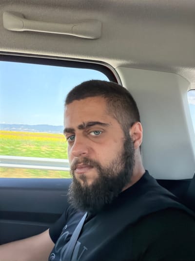 The image shows a close-up selfie of a man with a beard, looking directly at the camera. He is seated in the passenger seat of a vehicle, as indicated by the car's interior elements visible in the foreground, such as a grab handle on the ceiling and the headrest of the seat behind him. Through the window, a bright sunny sky and a landscape of yellow fields and distant hills can be seen, suggesting a rural or suburban setting. The man is wearing a black t-shirt and a grey seatbelt. The overall impression is that of a person taking a photo while traveling. Based on the provided context, the scene is located in the vicinity of Nedyalsko, Bulgaria, likely along a road traversing agricultural land.