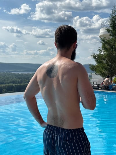The image depicts a man standing at the edge of an infinity pool, with his back to the camera. He has a dark beard and is wearing dark blue striped swim trunks. A dark circular tattoo is visible on his upper back, and his skin is speckled with water droplets. The pool's bright blue water reflects the sky, which is filled with scattered white clouds under a bright sky. In the background, a lush green forest stretches across rolling hills, with a body of water visible in the distance. To the right of the man, a large evergreen tree stands near a white, multi-level structure, possibly a building or observation deck. Further in the background, other people are visible relaxing on lounge chairs near the pool. The overall scene suggests a summer day at a resort or scenic overlook, likely in the outskirts of Novokuybyshevsk, Russia, given the natural landscape.