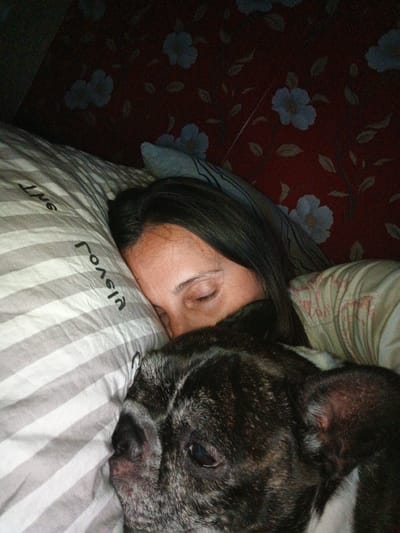 The image shows a close-up of a person and a dog sleeping in bed. The person's head is on a pillow with gray and white stripes. The person's face is partially visible, with their eyes closed. A dog, likely a French Bulldog, is also sleeping and is positioned in front of the person, with its head resting on the same pillow. The background appears to be a wall with a floral pattern in shades of red and light blue. The lighting is dim, suggesting it might be nighttime or the room is dark. The image does not contain any information that would suggest its location in the city of Goryachiy Klyuch, Russia.