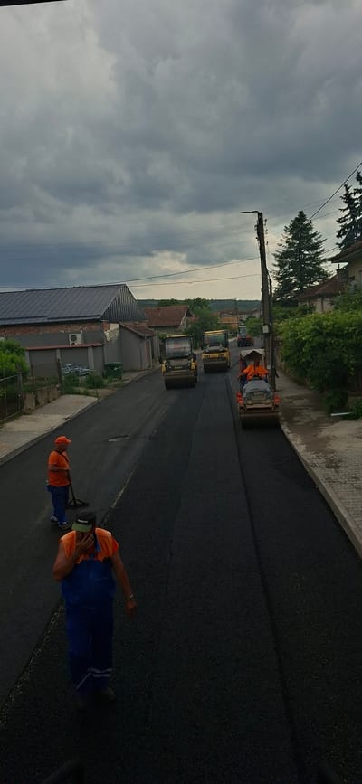 The image captures an active road construction scene in Strazhitsa, Bulgaria, under an overcast sky. The primary activity is the asphalting of a road, with multiple construction workers and heavy machinery involved.

Several workers are visible, dressed in orange high-visibility shirts and blue work pants, actively engaged in the process. One worker in the foreground is partially cropped, while others are seen further down the road, either operating machinery or overseeing the work. On one worker's cap, the text "FRONTIER" is visible.

The road itself is a prominent subject, covered in fresh, dark black asphalt which shows distinct lines from the paving and compacting process. Multiple road rollers or compactors are operating on the new surface; at least four can be identified. The brand name "HAMM" is partially visible on the side of one compactor, and "VOLVO" on another.

Along the side of the road, various residential buildings are visible with different roof types, including red tiles and grey metal. A prominent utility pole with numerous power lines runs parallel to the street. Lush green trees and bushes add to the surrounding vegetation. A concrete sidewalk or curb borders the asphalt. Additional objects include a green dumpster and some metal construction materials near one of the buildings.

The sky is heavily clouded with uniform grey tones, indicating a cloudy day. The scene depicts a typical infrastructure improvement project, showcasing the machinery and human effort involved in road maintenance or construction.