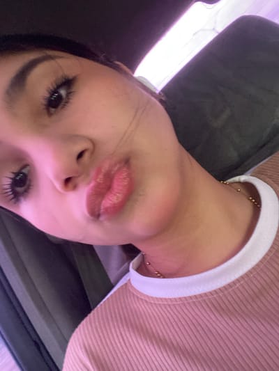 A close-up, rotated selfie of a person with light skin, brown eyes, and dark hair is presented. The individual is wearing a ribbed, light brown shirt with a white collar. A delicate gold necklace is visible at the base of the neck. The background is blurry and predominantly dark gray/black, appearing to be the interior of a vehicle. The setting cannot be confirmed to be El Dátil, Venezuela, from this image alone.