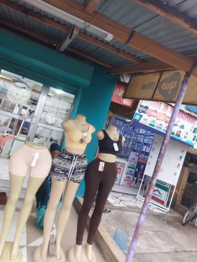 Here's a detailed description of the image:
**Setting:** A street scene, likely in a commercial area. There are structures resembling storefronts or businesses.
**Objects:**
* **Mannequins:** Three female mannequins. They are dressed in clothing and are posed horizontally.
* **Advertisements and Signage:** There are numerous signs and advertisements visible in the background. Brands like "NMB", "Tigo", "M-Pesa" are recognizable.
* **Building Structure:** Portions of a building are visible, including a roof structure with wooden beams and metal sheeting.
* **Assortment of items inside a glass door:** Clothing and textiles seem to be on display behind the mannequins.
* **Pipe or Rail:** There is a pipe or rail running horizontally across the image.
**Environment:**
* **Urban/Commercial:** The scene suggests a busy urban environment with commercial activities.
**Location Clues:**
* **Tanzania:** The presence of Swahili words (such as "Wakala" and "Mpesa") on the advertisements indicates the location is in Tanzania.
* **Dar es Salaam:** The brands like NMB, Tigo, and M-Pesa are commonly found in Dar es Salaam.