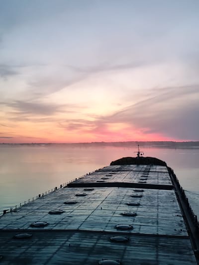 The image captures a freighter sailing on a body of water at sunrise. The sky is painted with hues of pink, orange, and purple, indicative of a colorful dawn. A gentle mist hovers over the water's surface. The freighter's deck, visible in the foreground, is dark and appears weathered, with rows of circular hatches. The stern of the ship, featuring a small structure with a mast and lights, is in the mid-ground. The far shore is lined with trees. The scene suggests a serene and tranquil morning commute on the water. The location is identified as Lukino, Russia.