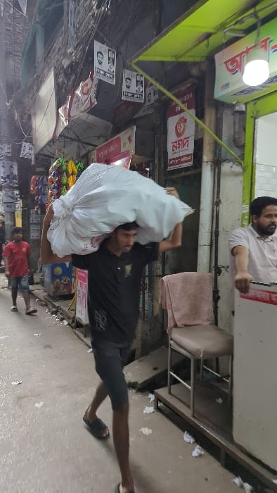 The image depicts a narrow, bustling commercial street or alleyway within an urban area of Dhaka, Bangladesh, under daylight conditions.

In the foreground, a young adult male, likely in his late teens or early twenties, with dark hair and a light beard, walks from right to left. He is wearing a black short-sleeved t-shirt with a white design, dark knee-length shorts, and black slide sandals. He carries a large, white, irregularly shaped sack on his left shoulder and head, supporting it with both hands.

In the mid-ground on the left, a second young male, wearing a red t-shirt and light denim shorts, walks away from the camera. On the right, a third male, with a beard and wearing a light-colored polo shirt, is partially visible, positioned at what appears to be a shop entrance, with his right hand resting on a white counter or appliance.

The street surface is paved and cluttered with scattered white litter, including crumpled papers and plastic. Buildings rise on both sides of the alley. On the right, a shop entrance features a prominent lime-green awning, a spherical white light fixture, and signs with Bengali script, notably the "Nagad" logo. A light-colored upholstered chair with a metal frame and a towel draped over it is placed on a raised platform outside this shop. On the left, another shopfront displays colorful packaged goods hanging for sale, and numerous political or advertising posters featuring faces and Bengali text are affixed to the building walls. A dense network of black electrical or communication cables crisscrosses overhead between the buildings.