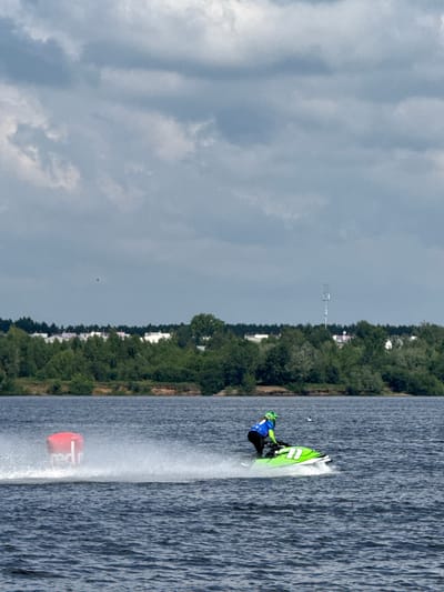 This is a photograph taken from a distance, showing a jet ski rider on a body of water, likely a river. The rider is wearing a blue and green racing suit and a helmet. They are on a green and white jet ski, which is kicking up a significant amount of spray as it moves. In the background, there is a shoreline with a dense line of green trees and some white buildings visible behind them. Further in the distance, a cell tower can be seen. The sky is overcast with dramatic, dark grey clouds. To the left of the jet ski, a red buoy with white lettering, appearing to say "red", is visible in the water, also creating spray. The water itself is dark and appears choppy. The general impression is of a water sport event taking place on a cloudy day. The presence of the cell tower and the style of the buildings suggest an urban or suburban environment. Given the mention of the city of Perm, Russia, it is probable that this scene is depicting a jet ski race or recreational activity on one of the city's waterways, such as the Kama River.