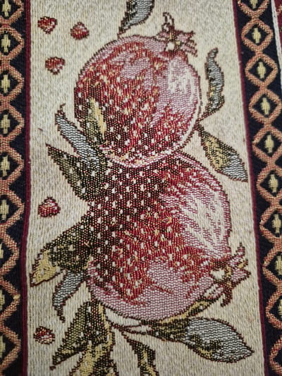 A close-up photograph captures a section of a woven textile, likely a tapestry or tablecloth, featuring two stylized pomegranates as its central motif. The pomegranates are depicted with intricate detailing, using a blend of red, dark red, and pinkish-white threads to create a textured appearance that suggests their internal arils. Leaves in shades of green, yellow, and black accompany the fruit. Several individual pomegranate arils are scattered across the light, neutral-toned background of the fabric.

The image prominently displays the woven texture of the textile, with individual threads and the weave pattern clearly visible, highlighting the craftsmanship. A decorative border frames the design at the top and bottom. This border features a repeating geometric pattern of interconnected diamond shapes, rendered in dark blue/black, reddish-brown, and golden-yellow threads, imparting a traditional aesthetic.

There is no explicit activity or event occurring; the image is a static artistic representation. The overall color palette is warm and earthy. Given the location context of Yerevan, Armenia, the pomegranate is a culturally significant symbol, often representing fertility, abundance, and prosperity in Armenian art and tradition. No visible text is present in the image.