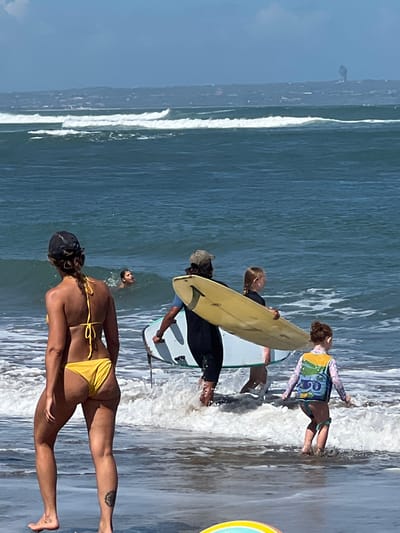 In North Kuta, Indonesia, on a sunny day, several people are at the beach with surfboards. In the foreground, a woman in a yellow bikini faces away from the camera, her back and legs visible. Her tattoos are prominent on her right thigh and left ankle. Behind her, a person in a black wetsuit and a person in a cap are near surfboards. To the right, a child wearing a rash guard and brown shorts stands in the shallow water, with a surfboard. Further out, a person's head is visible above the water. The ocean has white waves crashing on the shore. The background shows a distant shoreline with buildings and a hazy sky.
