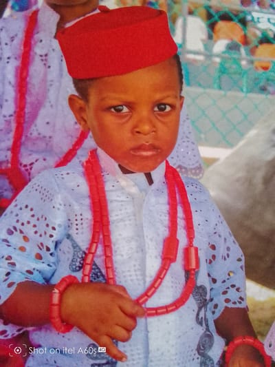 A young child is the primary subject of the image. The child wears a red fez hat, a white lace-patterned shirt, and multiple strands of red coral beads around their neck and on their wrist. The child stands, facing forward, and holds the coral beads in their hand. Another person is partially visible in the background to the left, also wearing a similar white lace-patterned garment and a red beaded necklace. The background shows a blurred outdoor scene. The text "Shot on itel A60s 4G" is in the bottom left corner, indicating the image was taken on that device. There is no indication that the image was taken in Idimu, Nigeria.
