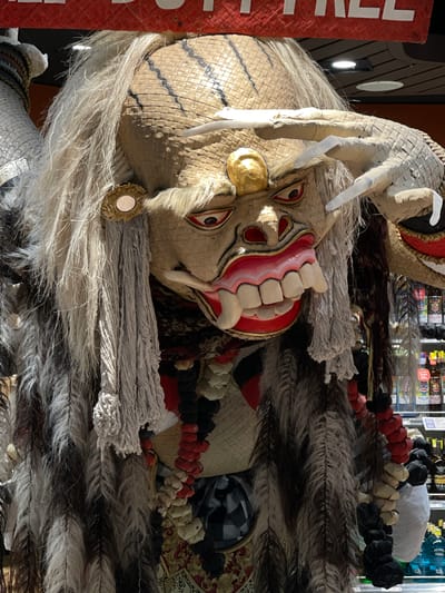 The image shows a close-up of a large, traditional Balinese mask, likely a Rangda mask, which is a prominent figure in Balinese mythology and dance. The mask is predominantly made of woven straw, with a light tan color forming the base. It features a fierce expression with large, red eyes outlined in black, and an open mouth revealing a row of sharp, white teeth and pink gums. The mask is adorned with a mane of long, white and grey hair that cascades down. There are also decorative elements such as red and white beads, and what appear to be straw or feather-like strands hanging from the sides. In the background, shelves with various bottles and products can be seen, suggesting the mask is displayed in a retail environment. A red banner with white text is partially visible at the top of the image. The setting appears to be a shop or market stall in Kuta, Indonesia, a popular tourist area known for its souvenir shops and cultural items.
