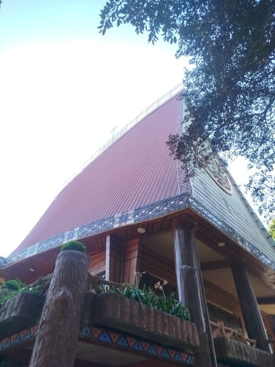 A low-angle shot of a building, possibly a church, located in An Phu Ward, Vietnam. The building features a high-pitched roof made of brown corrugated metal, decorated with a border of repeating geometric patterns. A cross is located at the top of the roofline. The building's facade includes a carved emblem and is supported by dark, cylindrical pillars. A decorative balcony with plant life and faux-wood pillars stands in the foreground. The sky is bright, with foliage from nearby trees framing the top of the image.