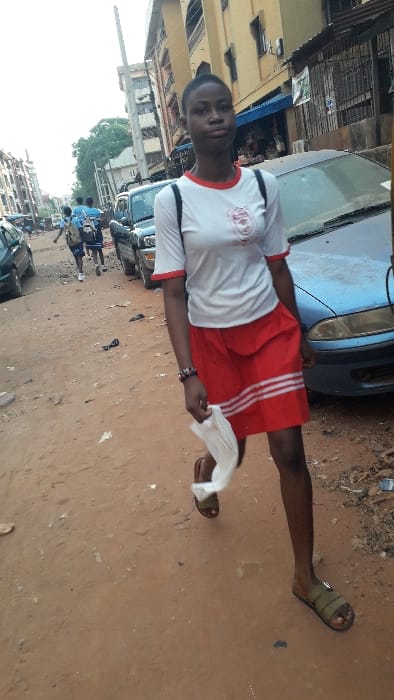 A young female, of dark complexion, walks on an unpaved, reddish-brown dirt street in Onitsha, Nigeria. She is positioned centrally, moving forward while looking towards the camera with a neutral expression. She wears a white short-sleeved t-shirt with red collar and cuffs, featuring a red-and-white crest on the left chest, and a red skirt with three horizontal white stripes near the hem. A black backpack is on her shoulders, a multi-colored bracelet on her right wrist, and olive-green slide sandals on her feet. She holds a crumpled white object in her left hand.
Immediately to her right is a parked, faded blue sedan. In the mid-ground, further down the street, several individuals, appearing to be students in blue shirts and carrying backpacks, walk away from the camera. The street is lined on both sides by multi-story concrete buildings, some with visible balconies and ground-floor shopfronts, including a prominent yellow/beige building on the right. Additional parked vehicles are visible along the street. A tall, light-colored utility pole stands in the mid-ground. The street surface is earthen with scattered debris. The sky above is bright and overcast.