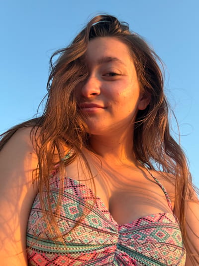 This image is a low-angle, close-up selfie of a young woman with long, light brown hair, taken outdoors against a clear blue sky. She is wearing a patterned bikini top with geometric designs in shades of pink, turquoise, and black. The woman is looking directly at the camera with a slight smile. The sunlight is hitting her face and upper body, casting a warm glow and creating some shadows. Her hair is windswept, with strands falling across her face. The bottom portion of the image clearly shows the texture and pattern of her bikini top, which appears to be ruched in the center. The background is uniformly blue sky, suggesting an open area without any visible structures or landmarks. The image quality is high, with good detail and natural lighting. The scene could be a beach or a similar outdoor recreational area. While the prompt mentions Sozopol, Bulgaria, there are no specific visual cues in the image to confirm or deny this location.