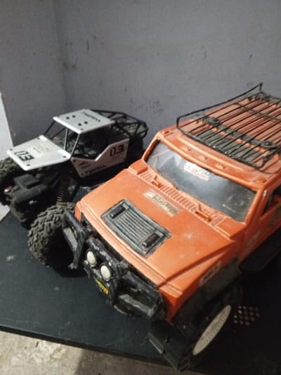 Two toy off-road vehicles rest on a dark surface. The larger, orange truck features a roof rack, a front grill with square indentations, and bumper lights. A sticker reading "SUPER ADVENTURE" is visible. Next to it, a smaller, white and black off-road vehicle with "03" printed on its hood and side displays a roll cage design. Both vehicles exhibit large, rugged tires suited for challenging terrain.