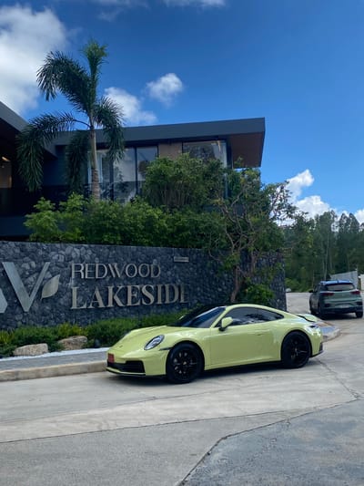 This media file shows a pale yellow Porsche parked on a paved driveway. The car is a two-door coupe with black rims. It is parked in front of a stone monument with "REDWOOD LAKESIDE" engraved in large, silver lettering. The monument also features a stylized "V" logo.
Behind the monument, there is a modern building with large windows and dark framing, suggesting a residential or commercial complex. Lush green trees and foliage surround the building and the monument. A palm tree is visible on the left side of the frame. In the background, more trees and a hillside are visible, hinting at a natural, possibly tropical setting. The sky is blue with scattered white clouds, indicating daytime with fair weather. Two other cars are parked further down the driveway in the background.
The overall scene appears to be the entrance to a luxury property or resort. The image is taken from a slightly elevated, horizontal perspective. No people are visible in the frame. The text "REDWOOD LAKESIDE" is clearly legible. The setting is consistent with Choeng Thale, Thailand, known for its upscale resorts and natural beauty.