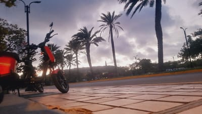 An outdoor, low-angle, wide shot shows a red electric scooter parked on a tiled sidewalk in the city of Pedro González, Venezuela. The scooter is positioned on the left side of the frame and faces towards the right. Behind the scooter, several palm trees are visible, silhouetted against a cloudy sky. The sky is dark and overcast, suggesting it is either early morning or late evening. In the distance, a lighthouse stands atop a hill. A road runs parallel to the sidewalk, with a yellow line marking its edge. Several cars are parked on the opposite side of the road. The scene is captured during what appears to be twilight.