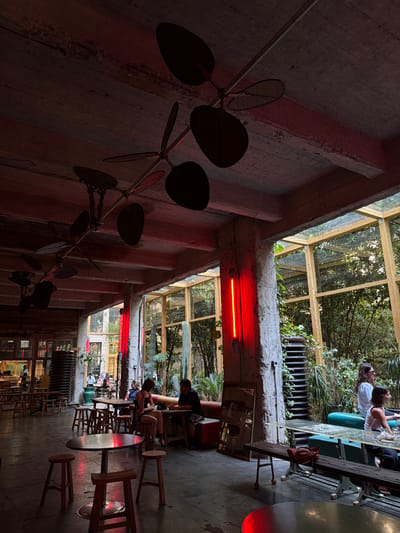The photograph captures the interior of a dimly lit, industrial-chic restaurant or bar, likely located in Tbilisi, Georgia, given the context of the overall scene. The space features exposed concrete ceilings and walls, with large, industrial-style ceiling fans suspended overhead. Red ambient lighting casts a warm glow across the scene, illuminating several areas and creating dramatic shadows.

The main area of the restaurant is furnished with numerous wooden stools and tables. In the foreground, a round table with four stools is centrally placed. Further back, other seating arrangements are visible, including a booth with upholstered seating and tables. People are seated at various tables, engaged in conversations or appearing to be dining.

The right side of the image opens up to a large glass-enclosed area, resembling a conservatory or atrium, which looks out onto a lush, green outdoor space with trees and plants. A tall, thin cactus-like plant is visible within this green space.

Distinctive red, vertical tube lights are mounted on a concrete pillar near the glass enclosure, adding to the establishment's unique lighting design. Stacked metal structures, possibly decorative elements or repurposed industrial items, are visible against the walls.

Several individuals are present. In the foreground, two people are seated at a table, a woman in a black top and a man. Further back, near the glass enclosure, two women are seated on a green sofa. In the background, more patrons are scattered throughout the space. The overall atmosphere suggests a vibrant and trendy gathering place.