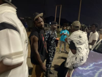The image shows a group of people, potentially a street gathering, in what is presumed to be Kubwa, Nigeria, given the prompt context. The lighting suggests it's nighttime.
People: Several individuals are visible, wearing various clothing styles. One is wearing a white and black patterned shirt, another a blue t-shirt, and at least one is in a black shirt. Two individuals appear to be wearing what looks like a Police uniform.
Objects: There are some poles visible. In the background, there appear to be automobiles.
Environment: The scene is at night. The ground is paved, possibly a street or open area.