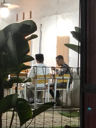 Two men are seated at a table on a patio at night in Lhokseumawe, Indonesia. The man on the left wears a white long-sleeved shirt and a black peci cap, and is facing away from the camera. The man on the right wears a camouflage-patterned shirt and is looking down at his food. They are seated at a white table with yellow accents. In the foreground, large dark green leaves obscure the view of the patio, and a metal fence is visible. The background wall is white, with three rectangular windows and vertical wooden planks. A ceiling fan is mounted on the ceiling.