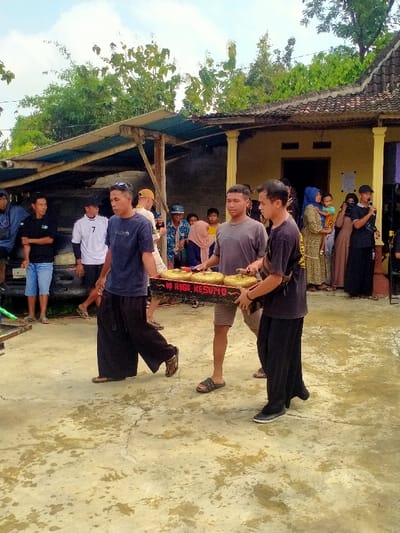 The scene is in front of a house in Kecamatan Kedawung, Indonesia, located at latitude -7.514, longitude 111.03. A group of men are gathered in front of the house. Three men are carrying a tray of golden musical instruments labeled "RI RAGO KESUMA." The men are dressed in casual clothing. Several other people are standing around, some watching the three men carry the instruments and some talking to each other. There is a car parked to the left of the men carrying the instruments. The scene is set in front of a house with a tan-colored facade. There are trees to the left of the house. The ground is covered with cement and there are trees in the background. The sky is partly cloudy.