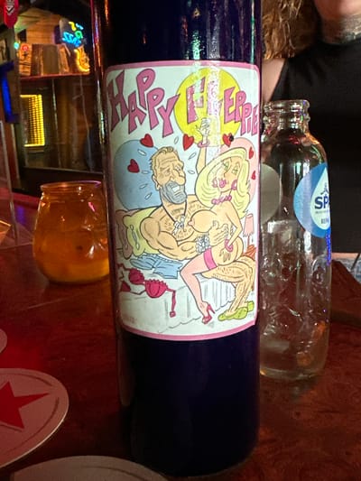 The image depicts a dark blue bottle with a cartoon label prominently displayed. The label features two cartoon characters, a man and a woman, in a suggestive pose, surrounded by pink hearts and text that reads "HAPPY ERRIE". To the right of the bottle is a clear glass bottle with a blue and white label that says "SPA REINE". In the background, partially visible, is a dimly lit establishment with neon signs and wooden paneling, suggesting a bar or restaurant setting. A small amber-colored glass jar is also present on the left. The overall scene appears to be indoors, likely in a bar or pub. There is no information in the image that suggests a specific location in The Hague, Netherlands.