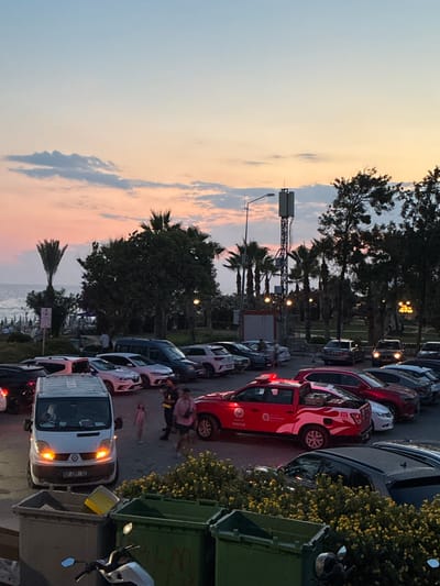 This is a vertically oriented, eye-level shot of a street in Alanya, Turkey, during sunset. The sky is filled with a gradient of soft blues, pinks, and oranges, indicating the time of day. Palm trees and other foliage are scattered throughout the scene, some casting long shadows. The street is lined with parked cars, including several red vehicles with distinctive flame graphics, and a white van with its headlights on. A large green dumpster and a motorcycle are also visible.
In the mid-ground, a group of people are gathered, one appearing to be lying down. Their presence suggests some sort of event or interaction is occurring. The overall mood of the image is a blend of everyday urban life and the serene beauty of a coastal sunset.
The visible text includes a license plate on the white van, "07 CFY 02". Additionally, a sign on one of the red cars reads "İTFAİYE", which is Turkish for "fire department".