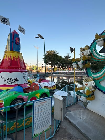 This image captures a children's amusement park ride in Đà Nẵng, Vietnam, likely located near the Han River or a similar waterfront area, given the calm water and reflections visible in the background. The ride features brightly colored, cartoonish cars designed to resemble race cars, with green, red, and white paint schemes. In the foreground, a green and pink car is prominently displayed, with a blue steering wheel and red seat. Behind it, other similar cars are parked.
To the left of the cars, a large, conical structure painted in yellow, red, blue, and white dominates the scene. It is decorated with flame-like patterns and topped with two checkered flags on poles, suggesting a racing theme. A sign with Vietnamese text, likely detailing rules and regulations for the ride, is attached to a blue fence in front of the cars.
To the right, part of another ride is visible, featuring animated figures that appear to be anthropomorphic cats or tigers, seated on a circular, teal-colored structure with green accents. One figure is clearly visible with yellow fur, white paws, and a green outfit. The background shows trees, buildings, and streetlights, indicating an urban setting. The sky is clear and blue, suggesting it's either early morning or late afternoon. The overall atmosphere is cheerful and designed for children's entertainment.