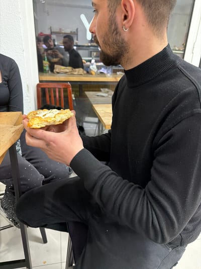 A man in a black sweater is holding a sandwich in his hands in a seated position indoors. The sandwich is filled with a white substance. The man has dark hair and a beard and is wearing an earring. There is a window behind him showing a reflection of people inside another space. The photograph was taken in Strumica, North Macedonia.