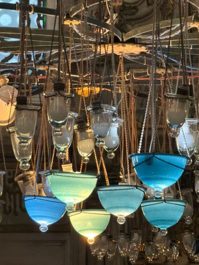 The image shows a close-up view of an ornate chandelier with multiple glass globes hanging from chains, likely located in the Eyüp Sultan Mosque complex in Eyüpsultan, Istanbul, Turkey. The chandelier features a variety of glass elements, including clear, blue, and greenish-blue globes of different shapes and sizes, some appearing to hold light sources. The chains are thin and metallic, creating a layered and intricate design. The background is dark and somewhat out of focus, suggesting an indoor environment, possibly a grand hall or prayer space within the mosque. The lighting in the image appears to be artificial, illuminating the glass elements and casting subtle reflections.