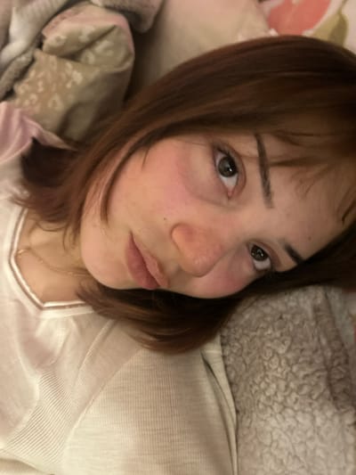 A close-up, slightly angled selfie captures a woman with light skin and shoulder-length brown hair. Her expression is soft, with a gentle pursing of her lips. She wears a light-colored, possibly white or cream, top with a simple neckline. A delicate gold necklace is visible. The background includes soft, textured bedding, suggesting a comfortable and relaxed setting. Her gaze is directed toward the camera, with dark eyes that appear tired. The lighting is soft, highlighting the contours of her face.