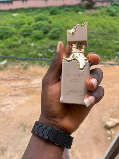 A person's hand, with dark skin and manicured nails, holds a beige and gold perfume bottle. The bottle has a unique, sculpted gold accent on the neck. Text on the bottle reads "ECLAIRE," "اكلاير," "EAU DE PARFUM," "VAPORISATEUR NATURAL SPRAY," "e50ml.," and "1.7 Fl. Oz." The person is wearing a black digital watch. The background is blurred, showing green foliage, suggesting an outdoor setting. The lighting indicates it is daytime. The image was taken in Gura Top, Nigeria.