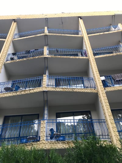The image shows a close-up, low-angle view of the balconies of a beige, multi-story building, likely a hotel or apartment complex, with blue metal railings. The balconies are stacked vertically, with each floor receding slightly from the one below. Some balconies have items of clothing hanging over the railings, suggesting they are occupied. The building's facade is textured, with a rough, stone-like finish on the horizontal elements. The lower portion of the image is partially obscured by lush green foliage, possibly a hedge or bushes. The sky is overcast and white, suggesting a cloudy day. Based on visual cues of the architecture and the commonality of such buildings in tourist areas, this scene is consistent with the Golden Sands resort area near Varna, Bulgaria, known for its hotels and vacation apartments. There are no people clearly visible in the image.