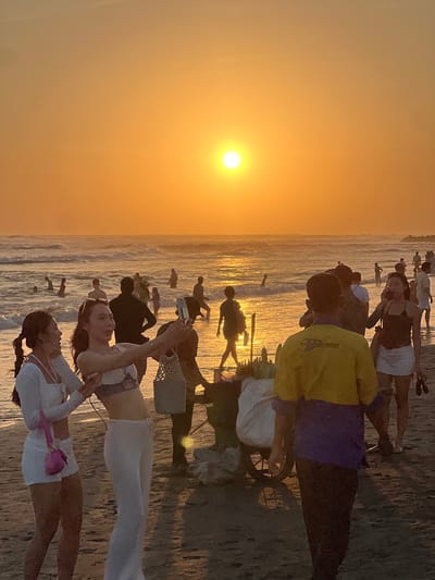 The image depicts a crowded beach scene at sunset in North Kuta, Indonesia. The sky is illuminated by a vibrant orange glow as the sun sets over the ocean, casting a warm light on the water and the sand. Numerous people are visible in the shallow water, wading and enjoying the waves. On the shore, several individuals are present, including two women in the foreground taking a selfie. One woman on the left wears a white top and shorts with a pink handbag, while the woman on the right is dressed in a white top and pants. Behind them, a vendor with a cart filled with corn is positioned near other beachgoers. The overall atmosphere is lively, with people engaged in various activities such as swimming, walking, and socializing.