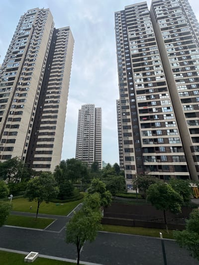 This image depicts a view of the Chongqing High-tech Zone in China, focusing on several tall residential buildings and a green courtyard. The buildings are modern, with numerous windows and balconies, and appear to be of similar architectural style. The courtyard features lush green grass, trees, and pathways, suggesting a well-maintained urban landscape. The sky is overcast, creating a soft, diffused light. No people are visible in the scene. The overall impression is one of a contemporary residential area within a city's technological hub.