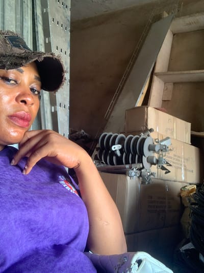 A close-up, high-angle shot depicts a person with dark skin lying down, partially visible. Their face, with glossy lips, is framed by a fringed cap. They are wearing a purple sweatshirt with visible lettering. Behind them are boxes and what appears to be electrical equipment. The setting seems to be an interior space with shelving and various objects stored, which is located in the city of Shaba, Nigeria.
