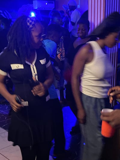 This image depicts a scene of revelry within a dimly lit, indoor venue in Durumi, Nigeria. The atmosphere is charged with blue lighting, reflecting off the dark, polished floor and the striped, vertical wall decor. In the foreground, a woman with long, dark hair adorned with a checkered headband is captured mid-dance, wearing a black dress with white trim and metallic buttons. Her right hand is raised, holding a dark object, possibly a phone. To her right, another woman, her face obscured, is also dancing, clad in a white tank top and blue pants. Her left hand is raised, holding a bright orange cup. In the background, other individuals are visible, some dancing, others observing the scene. One person in the mid-ground wears a blue shirt and appears to be dancing. The overall impression is one of a lively social gathering, possibly a party or a club night.