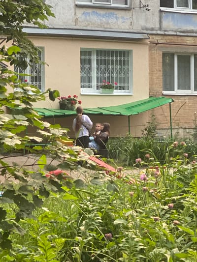 In Poltava, Ukraine, two young girls are seated on a slide under a green awning. One girl, wearing a dark top and blue jeans, holds a light-colored object, possibly a toy. The other girl, wearing a white hoodie and black pants, stands behind her. They are situated in front of a tan building with barred windows, where potted red flowers are visible on the windowsill. In the foreground, lush green foliage and pink flowers obscure parts of the scene. The background consists of a brick building and a grey, weathered facade.