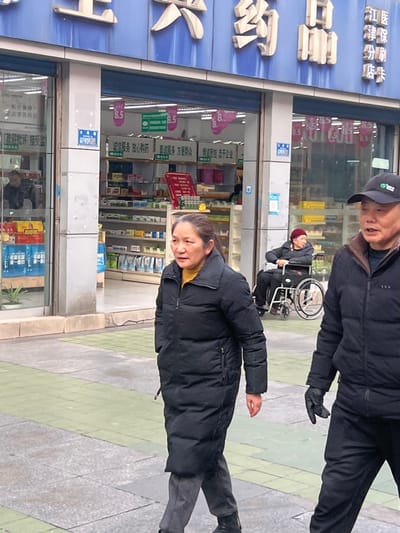 The image depicts a street scene in Jiangjin, China, focusing on the entrance of a pharmacy identified by its prominent blue facade with white Chinese characters reading "大兴药品" (Daxing Pharmaceutical) and a partial sign indicating "江津医保分店" (Jiangjin Medical Insurance Branch Store). Inside the well-lit store, numerous packaged goods are visible on shelves, along with several signs displaying "8.5" and Chinese text, including "诚信服务 放心购药" (Honest Service, Rest Assured to Buy Medicine).

In the foreground, an East Asian female, middle-aged to elderly, with dark hair tied back, walks from right to left. She wears a long black puffer coat over a yellow top, grey trousers, and black shoes. Her right hand is in her coat pocket. To her right, partially visible, is an East Asian male, middle-aged to elderly, wearing a black baseball cap, a black puffer jacket, dark trousers, and a black glove on his visible left hand, also walking from right to left.

In the midground, positioned just inside the pharmacy's open entrance, an elderly East Asian individual wearing a dark jacket and a red knit cap sits stationary in a manual wheelchair. The ground is a paved sidewalk with alternating grey and green square tiles. All visible individuals are dressed in winter attire, suggesting cool or cold weather conditions.