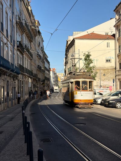 A tram travels down a narrow, steep street in Lisbon, Portugal. The tram is yellow and white with "THE ART OF SHOPPING" written on its side. Several people are visible inside the tram. The street is lined with tall, historic buildings with many windows and balconies. The sky is clear and blue, suggesting a sunny day. The sun casts long shadows, indicating it is likely late afternoon. Several people are also visible on the sidewalks, some sitting on steps and others walking. Tram tracks run down the center of the street. A black car and a white van are parked or driving further up the street. The overall scene depicts a bustling urban environment in a European city.