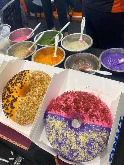 The image depicts a close-up, eye-level view of two donuts in white cardboard boxes, presented at a stall or event. In the background, several metal bowls are visible, each containing a different colored frosting and a spatula: pink, green, tan, orange, brown, and purple. These bowls appear to be set up for decorating donuts. The donuts themselves are prominently displayed. The donut on the left is half-covered in orange frosting with chocolate chips and half in tan frosting with small, crunchy sprinkles. The donut on the right is half-covered in pink frosting with red crumble topping and half in purple frosting with yellow crumble topping. The overall scene suggests a dessert-making or tasting event, potentially a pop-up shop or a booth at a fair. The lighting is bright and focused on the donuts. The backdrop is blurred but suggests an indoor setting with hints of people and furniture. The specific location of Davao City, Philippines is not discernible from the visual information provided in the image.