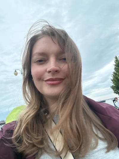 This is a selfie taken by a young woman with long, blonde hair. She is wearing a dark red jacket over a gray shirt. Her face is partially obscured by her hair, but she appears to be smiling. The background is a cloudy sky and some trees, suggesting an outdoor setting. In the lower part of the image, a street lamp and a portion of a bright yellow object are visible. The overall mood seems casual and relaxed. The image appears to have been taken during the day, with overcast weather. There is no visible text in the image.