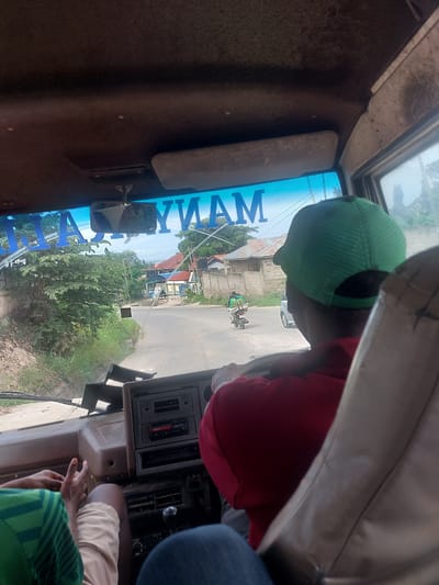 The image, taken from inside a vehicle, shows a view of a street in what is likely a residential area of Dar es-Salaam, Tanzania. The name "Mamyam" is visible, potentially indicating a business name or route identifier. A person wearing a green cap and red shirt is in the front seat. Through the windshield, one can see buildings, vegetation, and a road with a motorcycle and possibly a car. The vehicle's dashboard is visible, along with other passengers.