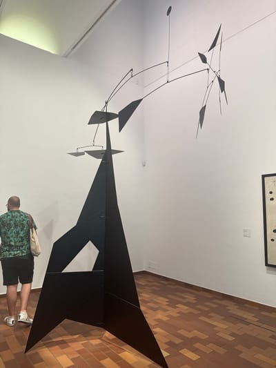 An outdoor art exhibit in Barcelona, Spain, features a large, black, abstract sculpture with multiple geometric shapes. The sculpture is composed of sharp, angular planes, with some elements appearing to be suspended in the air. A man with a bald head, wearing a green and blue floral print t-shirt and black shorts, stands to the left of the sculpture, facing away from the camera. He is carrying a light-colored tote bag over his left shoulder. The floor is paved with terra cotta tiles arranged in a staggered pattern. In the background, a white wall displays a framed artwork with black dots and lines on a light background, and a smaller information plaque. A light fixture is visible at the top of the wall, casting a pale green glow.