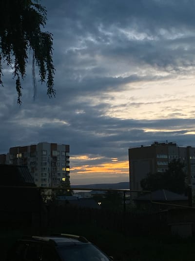 This image depicts a twilight scene in the city of Votkinsk, Russia, likely taken from an elevated position looking towards the west. The sky is dominated by dramatic, dark clouds with streaks of orange and yellow light breaking through near the horizon, indicating the setting sun. In the mid-ground, two apartment buildings are visible. The building on the left is lighter in color with orange accents and illuminated windows. The building on the right is darker and appears to be a residential block with many windows. Between and below these buildings, a darker silhouette of trees, smaller houses, and possibly a body of water can be seen in the distance. In the foreground, the dark shape of a car's roof and a section of a dark structure, possibly a fence or another building, are visible. A branch of a tree with long, thin leaves hangs from the upper left corner of the frame, partially obscuring the sky. The overall atmosphere is somber and peaceful.