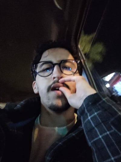 A person wearing glasses lies on their side, a hand touching their mouth. The individual has dark hair and a light-colored complexion. They are wearing a light blue undershirt and a dark plaid shirt. The background is mostly dark. A window with a reflection is in the foreground, and some lights are visible through the window. The image appears to be taken in a car at night.