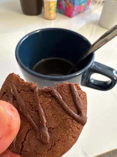 In a close-up, overhead shot, a finger is holding a chocolate cookie with a bite taken out of it. The cookie is dark brown with a drizzle of chocolate icing and has a cracked texture. Behind the cookie, a dark blue mug with coffee and a spoon is visible. The setting appears to be a table, with a blurred background suggesting a domestic or casual environment. The image does not provide any specific details that would indicate its location in Kuala Lumpur, Malaysia.